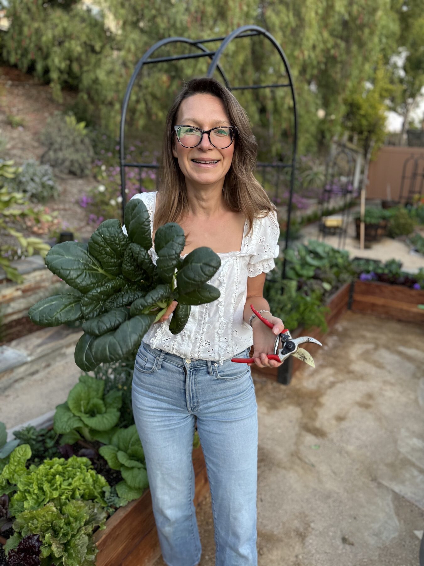 Marynika smiling, holding freshly harvested greens beside a garden arch trellis